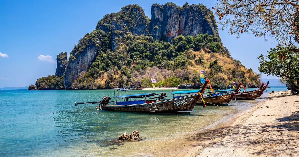 Longtail boats moored on a bright white-sand beach in southern Thailand beneath limestone rock formations, the kind of trip travellers now plan around the 30-day visa-exemption rule of 15 September 2026
